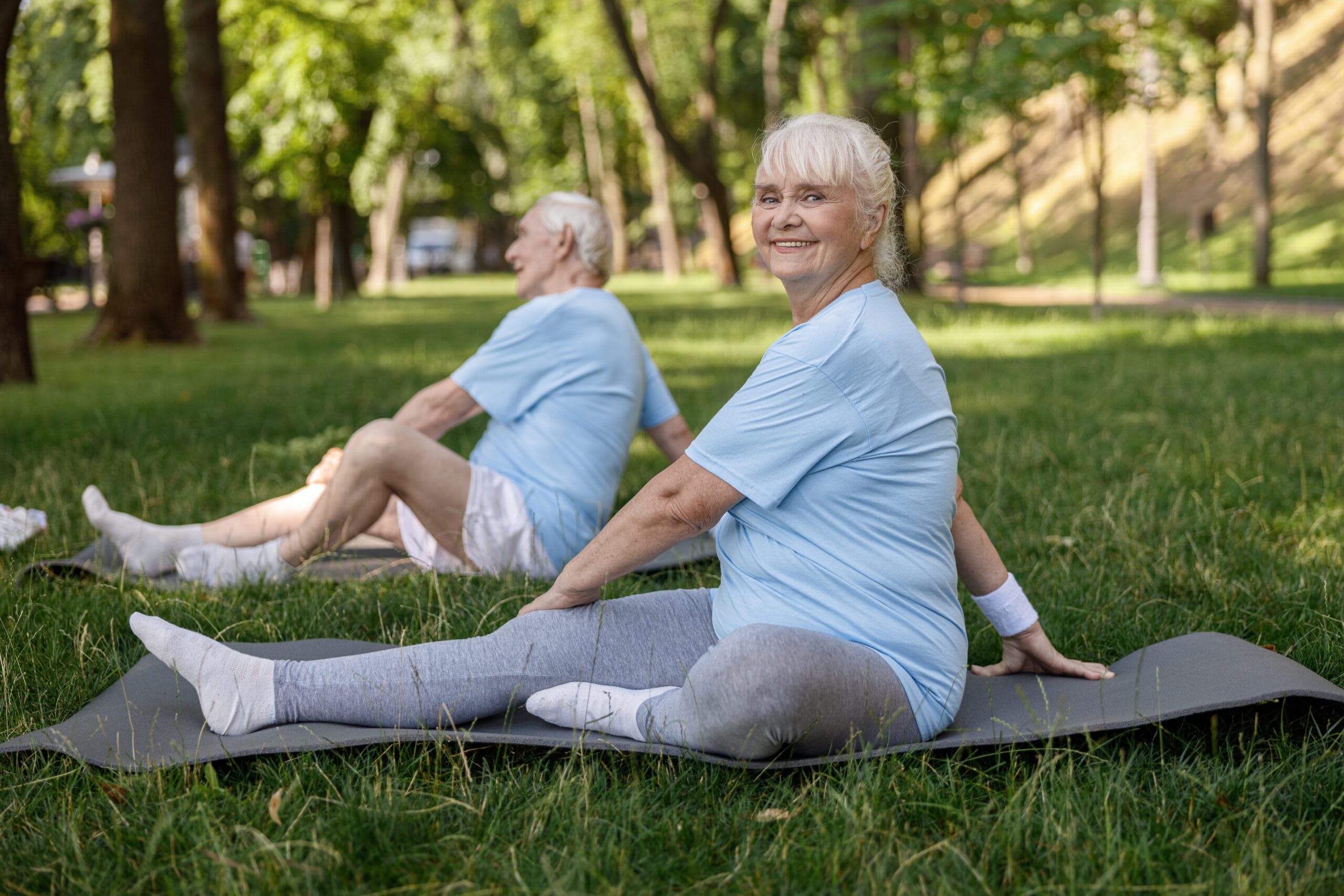 Two seniors stretch on a yoga mat in the grass.