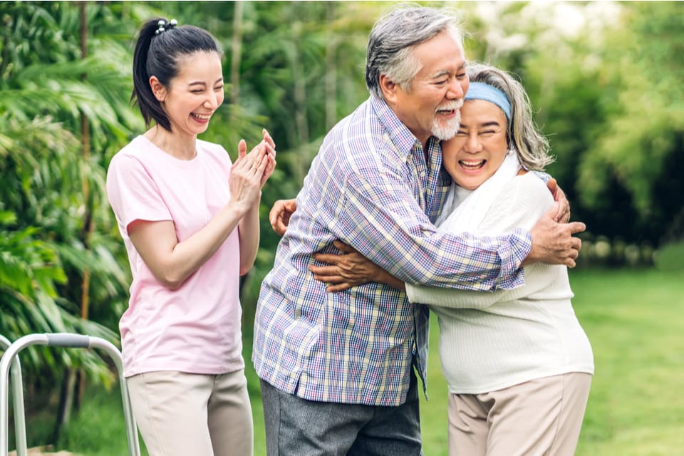 A senior man and woman hug while a young woman claps.