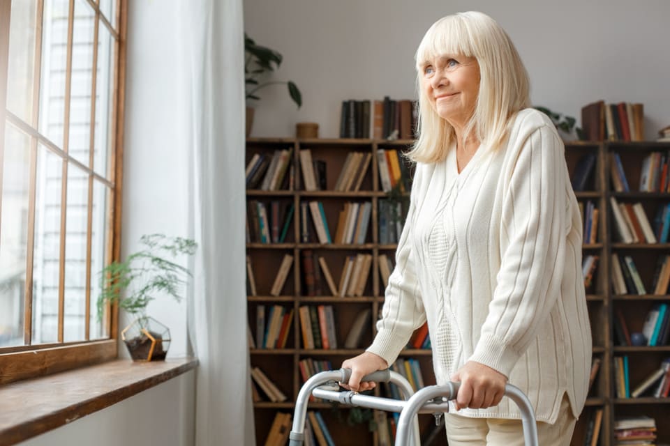 A senior woman uses a walker while staring out a window.